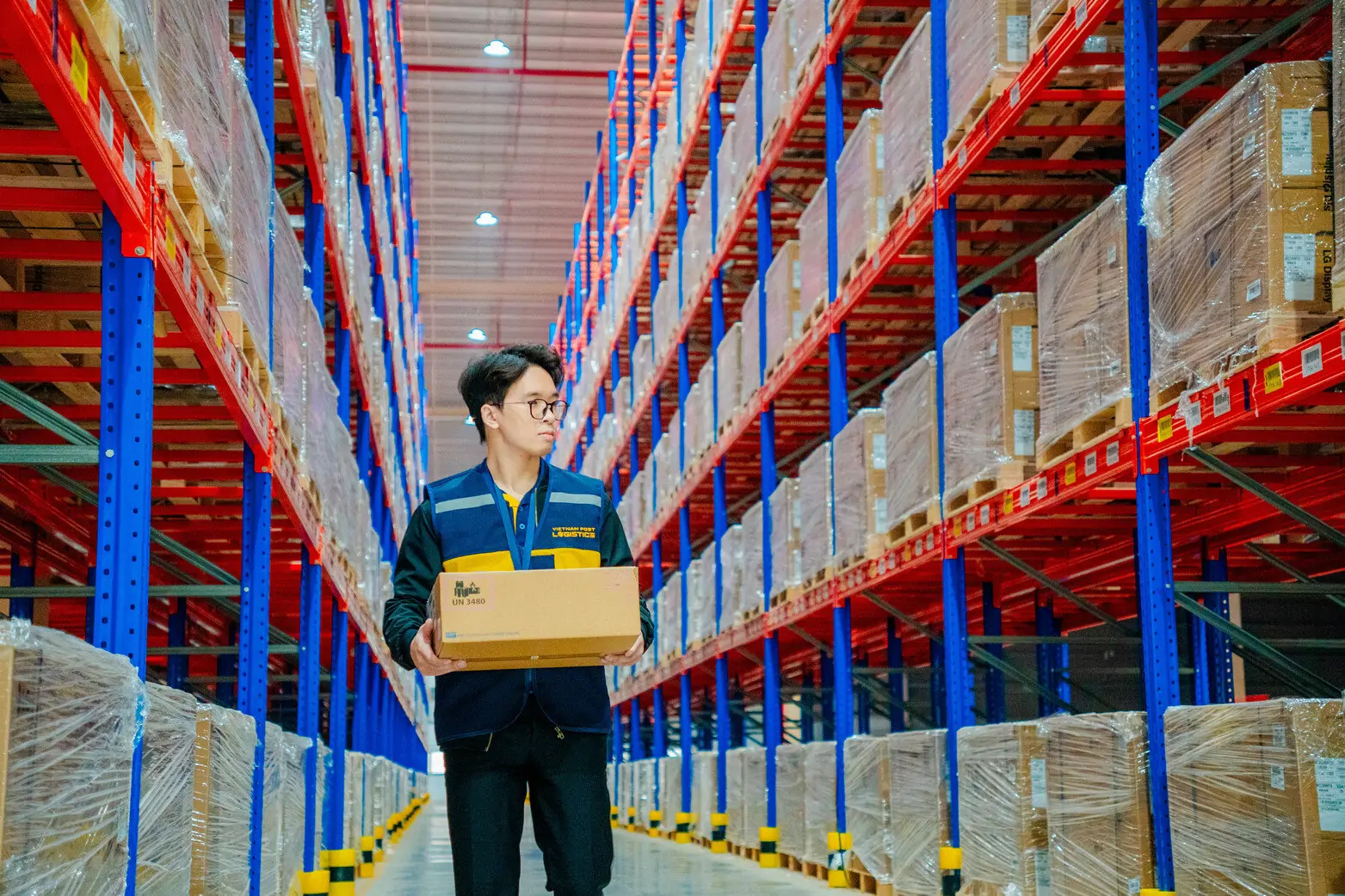 Nhân viên đang cầm thùng hàng trong kho, xung quanh là các kệ hàng hóa được sắp xếp gọn gàng.
